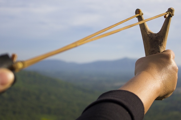 A hand holding a slingshot aims at an unseen target with a mountainous landscape in the background