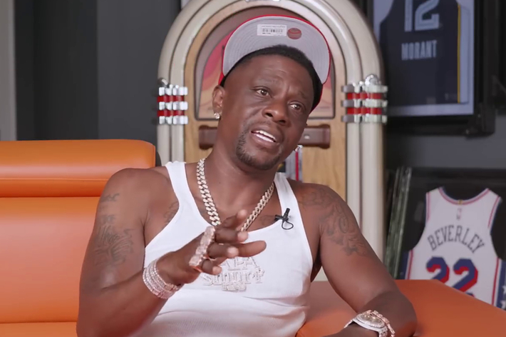 Boosie Badazz sits in an orange chair, wearing a white tank top, cap, and multiple chains, gesturing while speaking. A jukebox is behind him