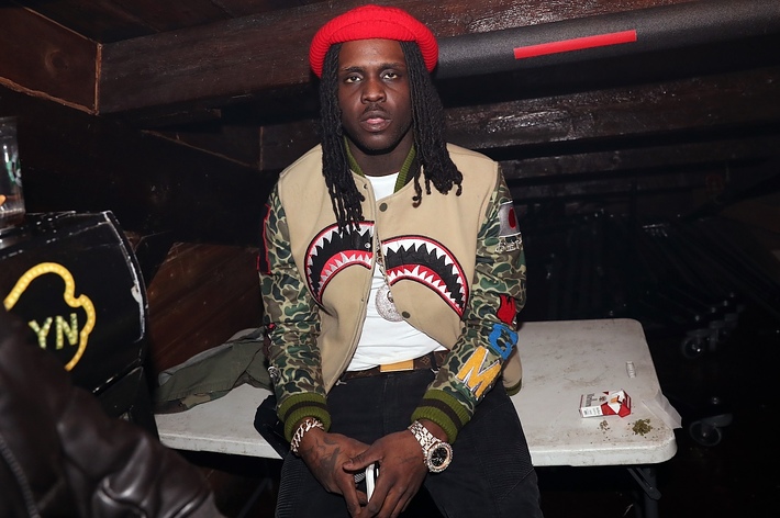 Man seated wearing a red beanie, camo jacket with shark mouth design, and jewelry