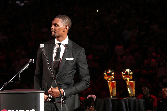 Chris Bosh stands at a podium in a suit during an event. Two NBA championship trophies are displayed on a table beside him