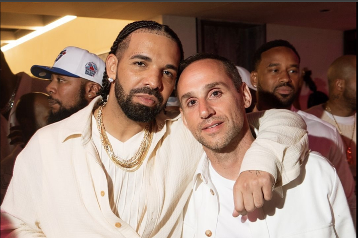 Drake and another man at a social event, both dressed in casual white shirts. Drake has his arm around the other man's shoulder while holding a drink