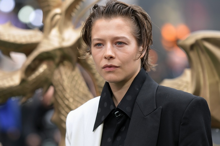 Emma D’Arcy at a public event wearing a half-white, half-black blazer with a polka dot shirt, standing in front of a decorative dragon sculpture