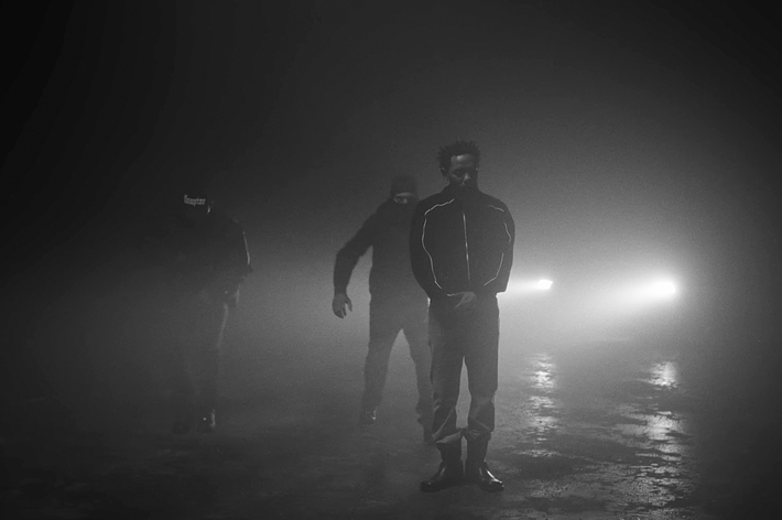 Three individuals, including a man in a track jacket, standing in a foggy, dimly-lit environment with headlights in the background