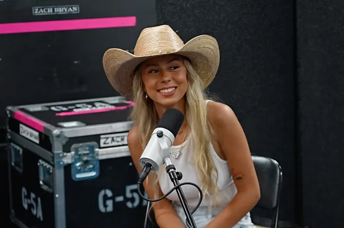 A woman in a straw cowboy hat and casual clothing smiles while sitting at a microphone during a recording session. A "Zach Bryan" sign is visible in the background