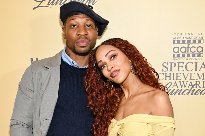 Two people posing at an event, one with a hat and blazer, the other wearing an off-shoulder dress