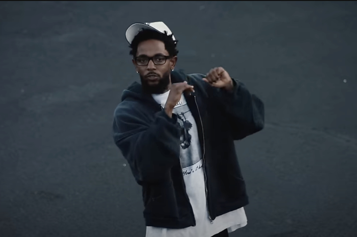 Kendrick Lamar stands outdoors, wearing a dark jacket, graphic tee, and baseball cap, gesturing with his hands