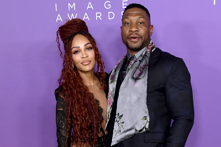 Two individuals on a purple backdrop; the person on the right sports a floral scarf and suit