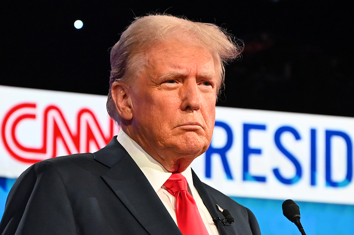 Donald Trump stands in front of a backdrop with the CNN logo and the word "President." He is wearing a suit with a white shirt and red tie