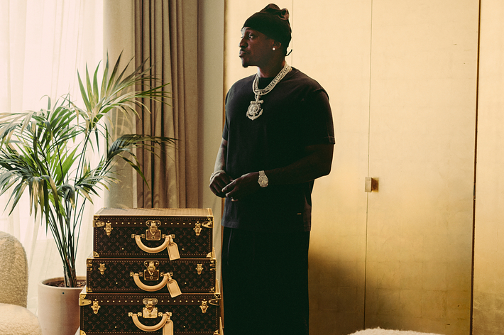 Person stands indoors near stacked designer luggage, wearing a black t-shirt, black hat, and large jewelry in a stylish, relaxed pose