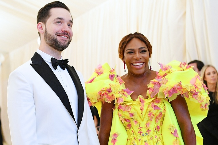 Alexis Ohanian and Serena Williams on a red carpet. Ohanian wears a black and white tuxedo; Williams in a vibrant dress with ruffled shoulders and floral designs