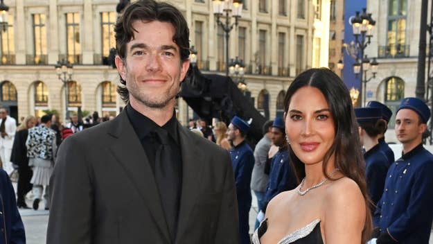 John Mulaney in a black suit and Olivia Munn in an elegant gown at a formal event, standing outdoors