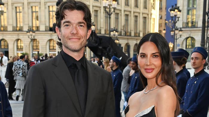 John Mulaney in a black suit and Olivia Munn in an elegant gown at a formal event, standing outdoors