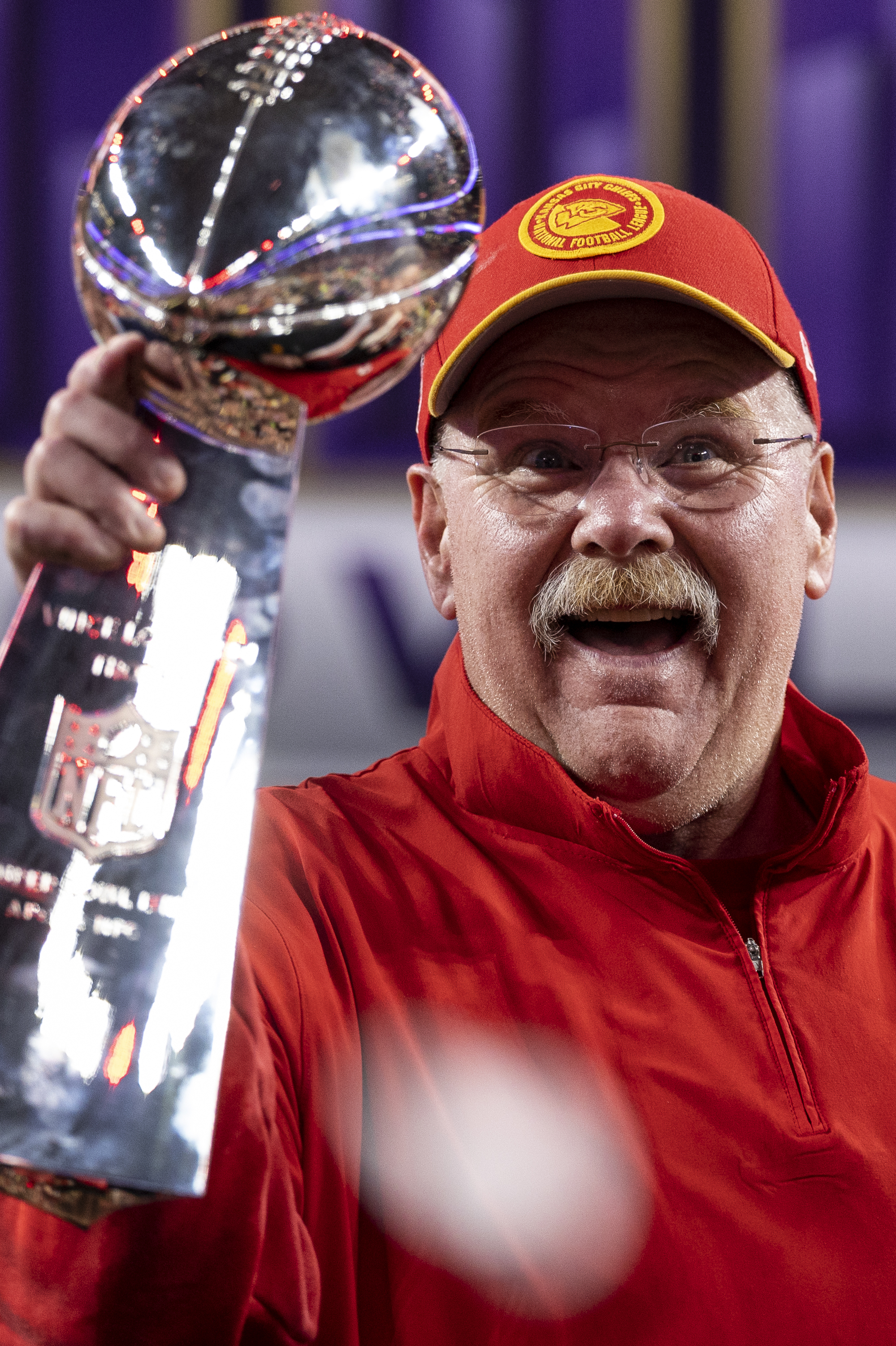 Andy Reid, in a jacket and cap, holds up the Vince Lombardi Trophy with a joyful expression on his face