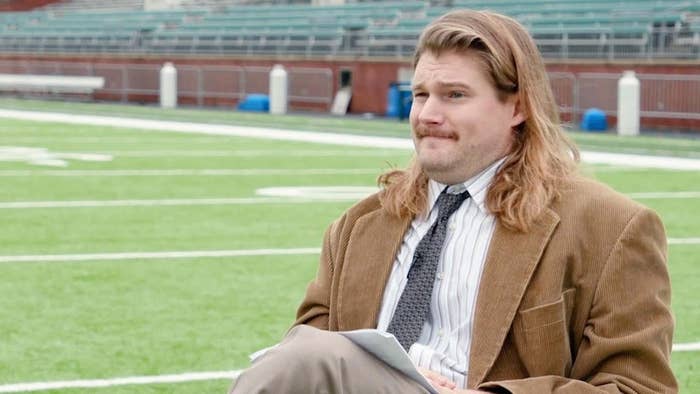 Man with long hair and mustache in brown suit, seated on a football field, holding paper