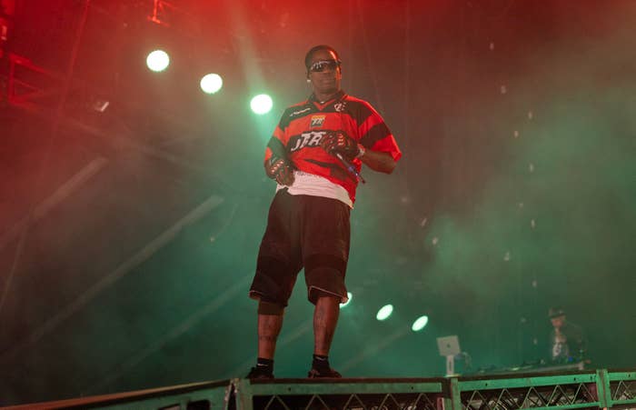 Travis Scott performs on stage, wearing a jersey, shorts, and sports glasses. Stage lights and smoke enhance the atmosphere