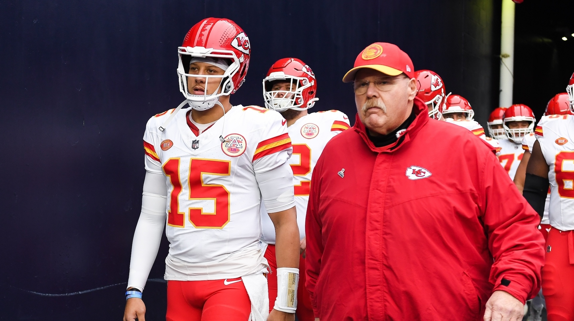 Patrick Mahomes and Andy Reid, dressed in Kansas City Chiefs attire, walk together, surrounded by team members. Mahomes wears a football uniform, and Reid wears a jacket and cap