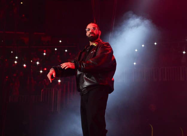 Drake performing on stage, wearing a leather jacket and black pants, with a misty background and audience lights in a concert arena