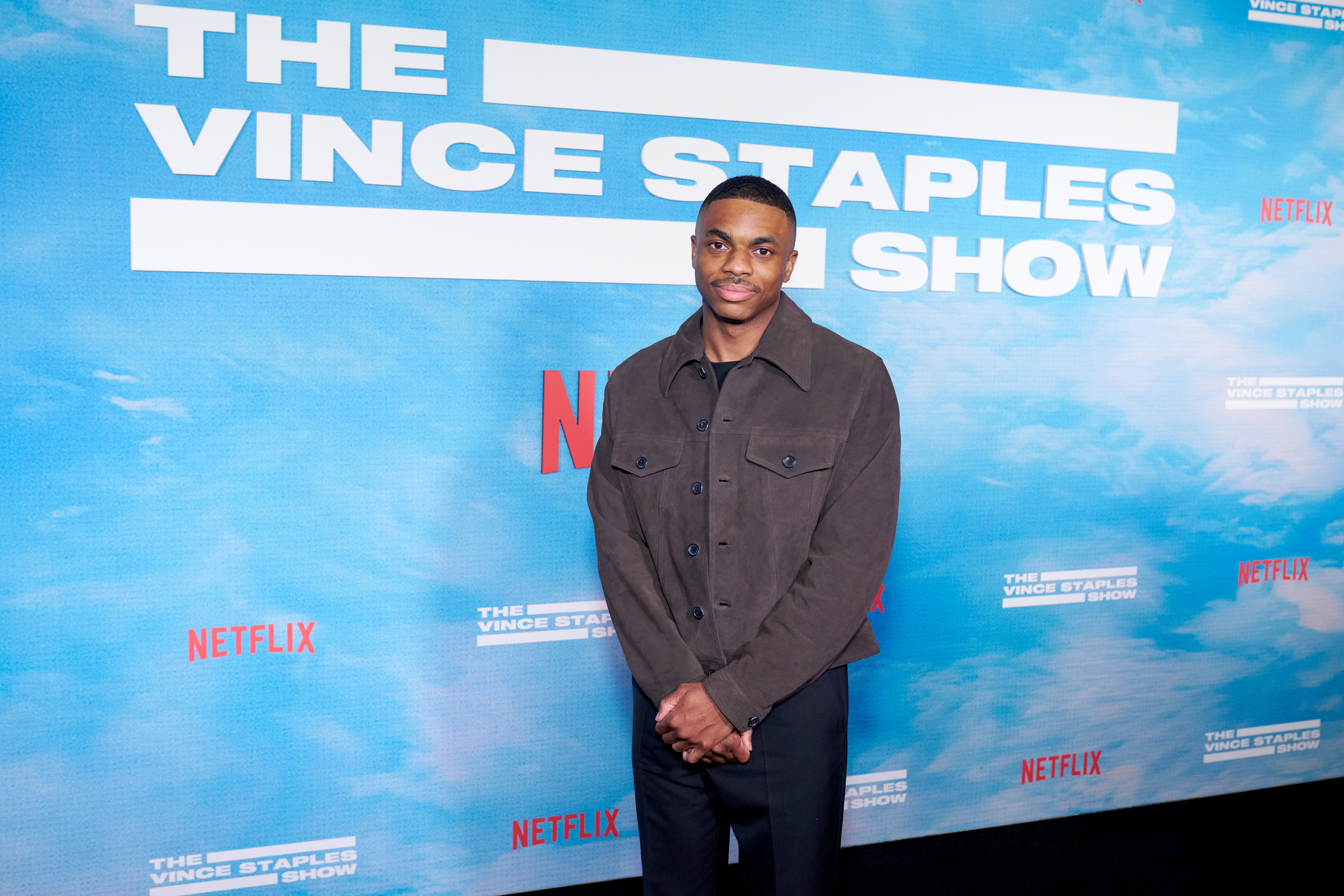 Vince Staples is standing in front of a backdrop that reads &quot;The Vince Staples Show&quot; at a Netflix event. He is wearing a dark jacket and pants