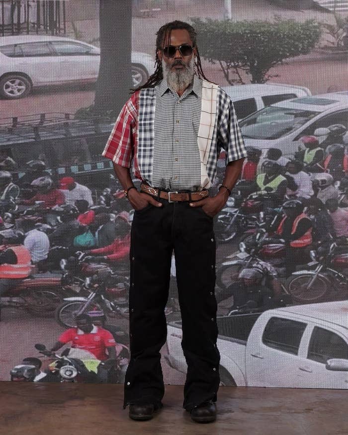 Man in a patchwork plaid shirt, sunglasses, and black pants with his hands in his pockets, standing in front of a busy urban street backdrop