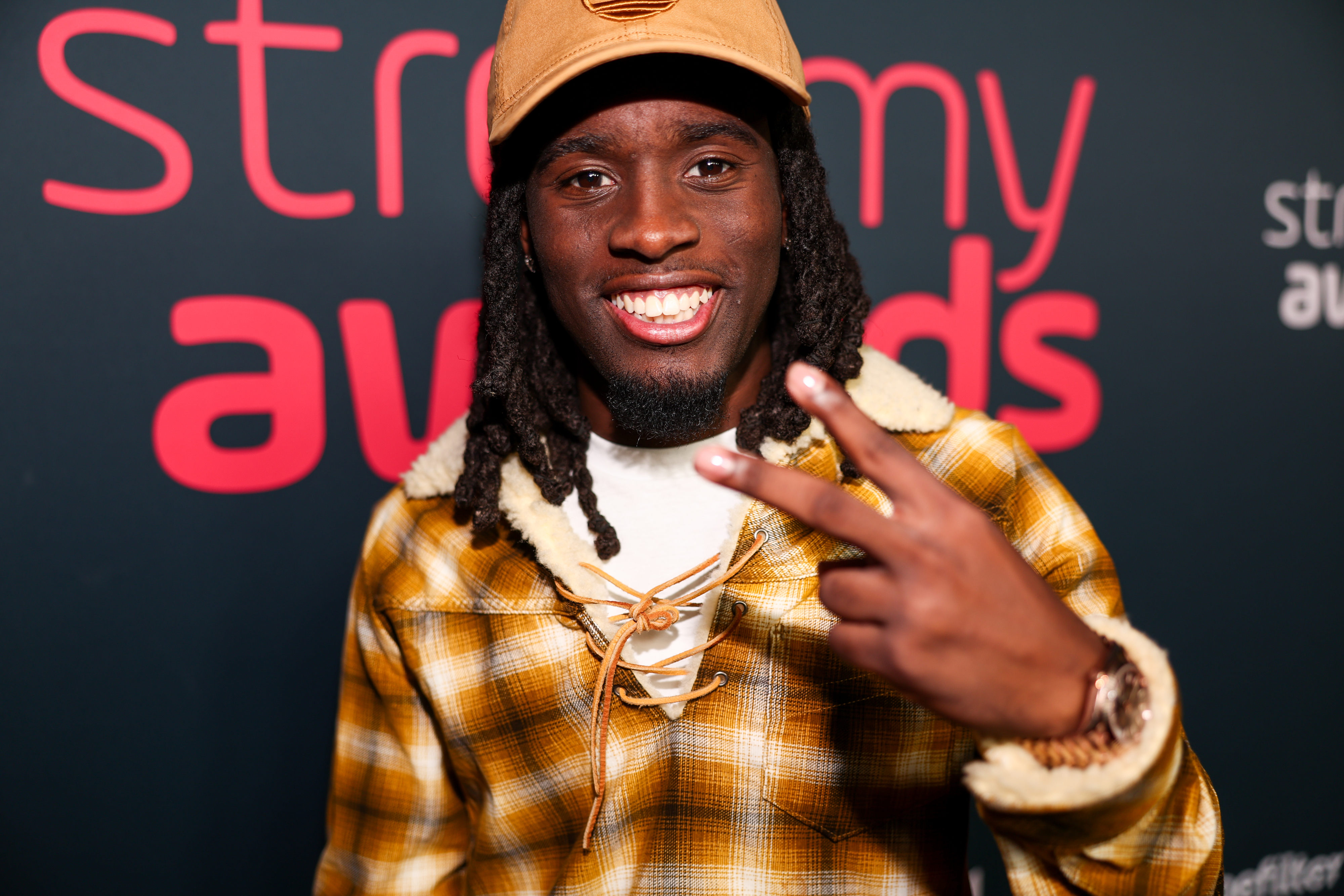 Kai Cenat at the Streamy Awards, wearing a plaid jacket with shearling collar and a tan cap, smiling and holding up a peace sign