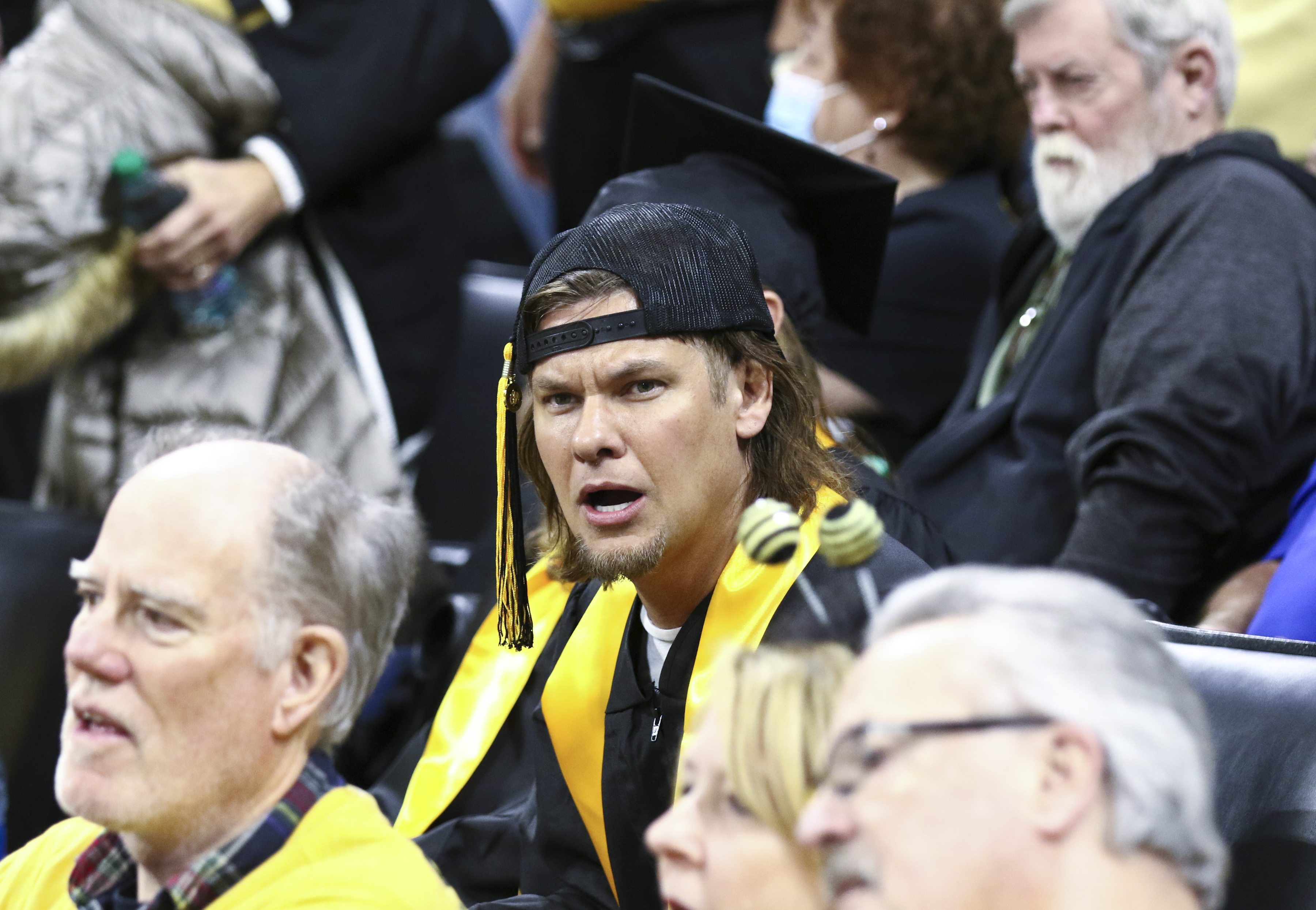 Theo Von, wearing a graduation gown and cap, sits among a crowd. He looks surprised or confused