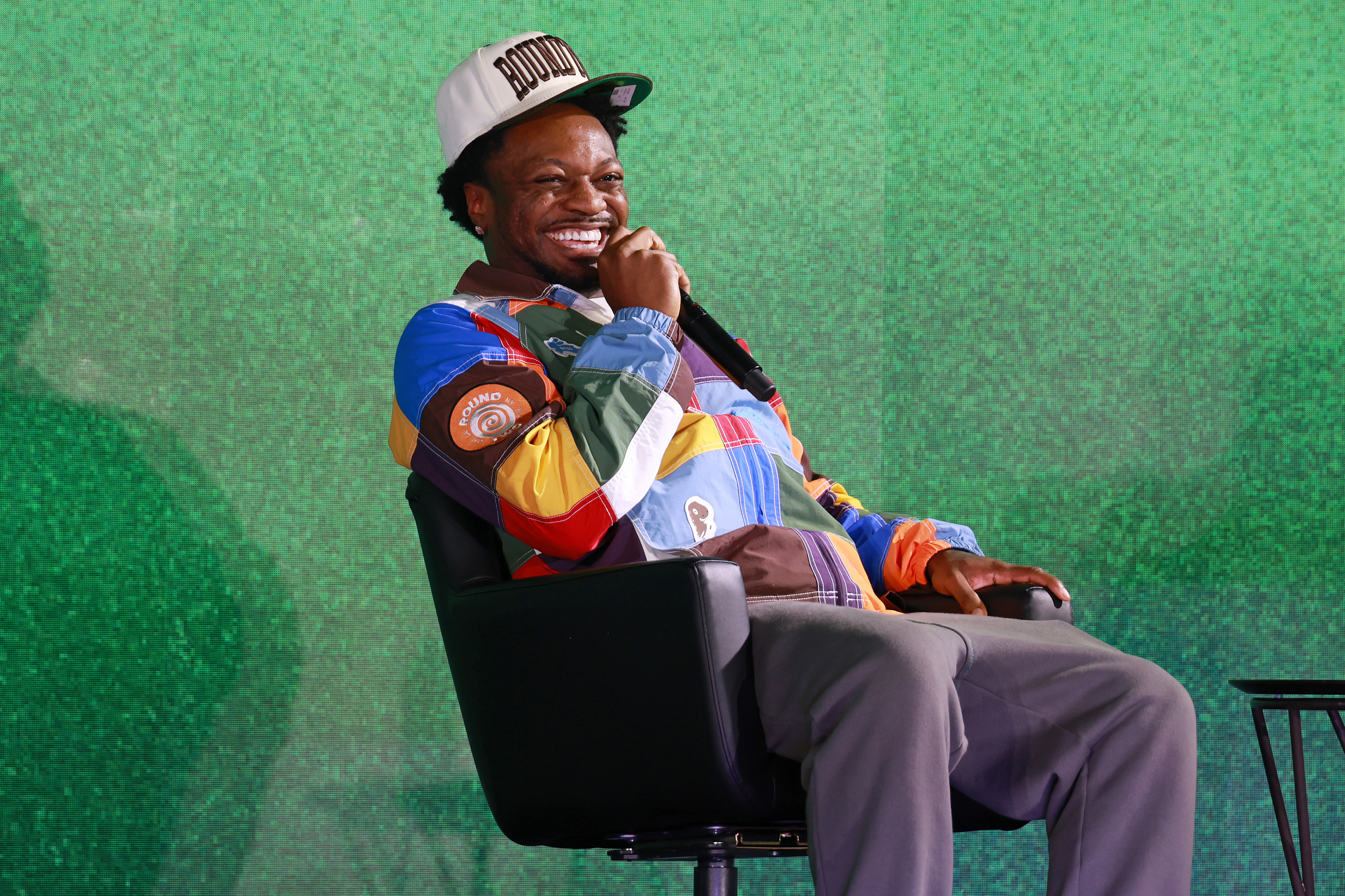 Jermaine Fowler speaks into a microphone while seated, wearing a colorful jacket and a white cap