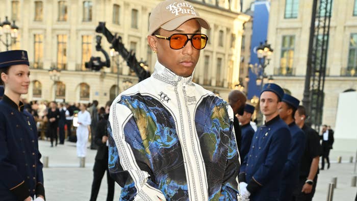 Pharrell Williams stands outdoors at a public event, wearing an ornate jacket, a khaki cap, and orange-tinted glasses. Uniformed staff are visible behind him