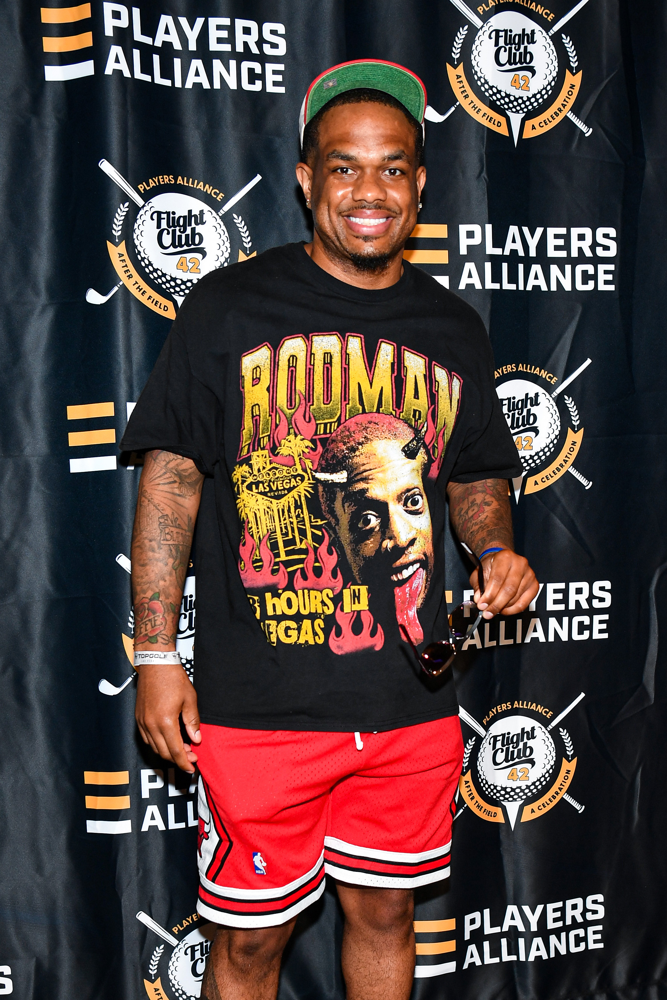 Man in a Rodman shirt and red shorts at Players Alliance event, smiling, holding sunglasses, standing in front of a backdrop with logos