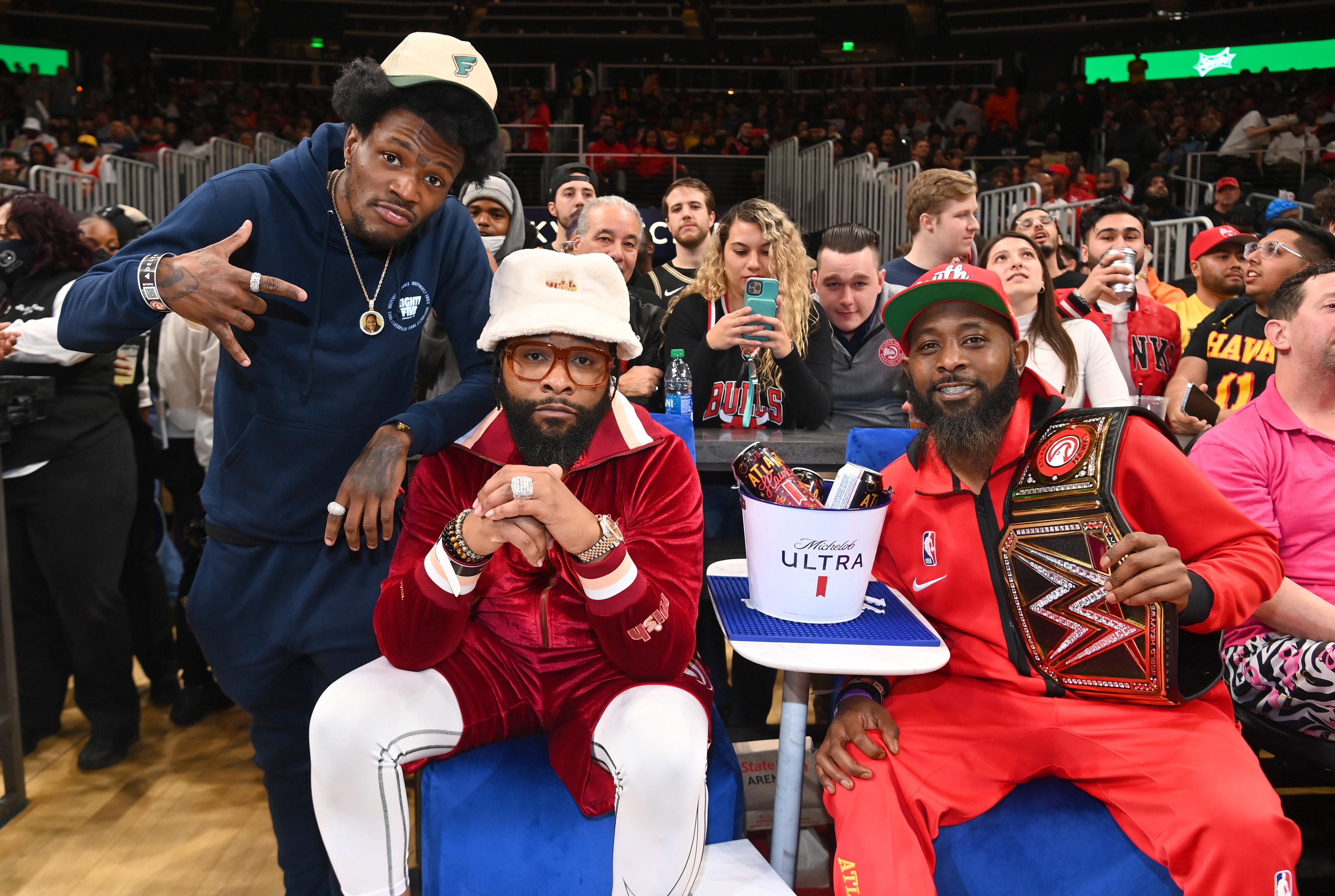 Devin Booker, Jalen Rose, and James Harden sit courtside at a basketball game. Rose wears a red tracksuit, Booker sports a casual outfit, and Harden holds a championship belt