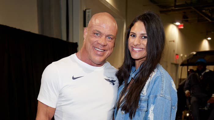 Kurt Angle, wearing a fitted white athletic top, poses with Giovanna Yannotti, dressed in a casual denim jacket, indoors