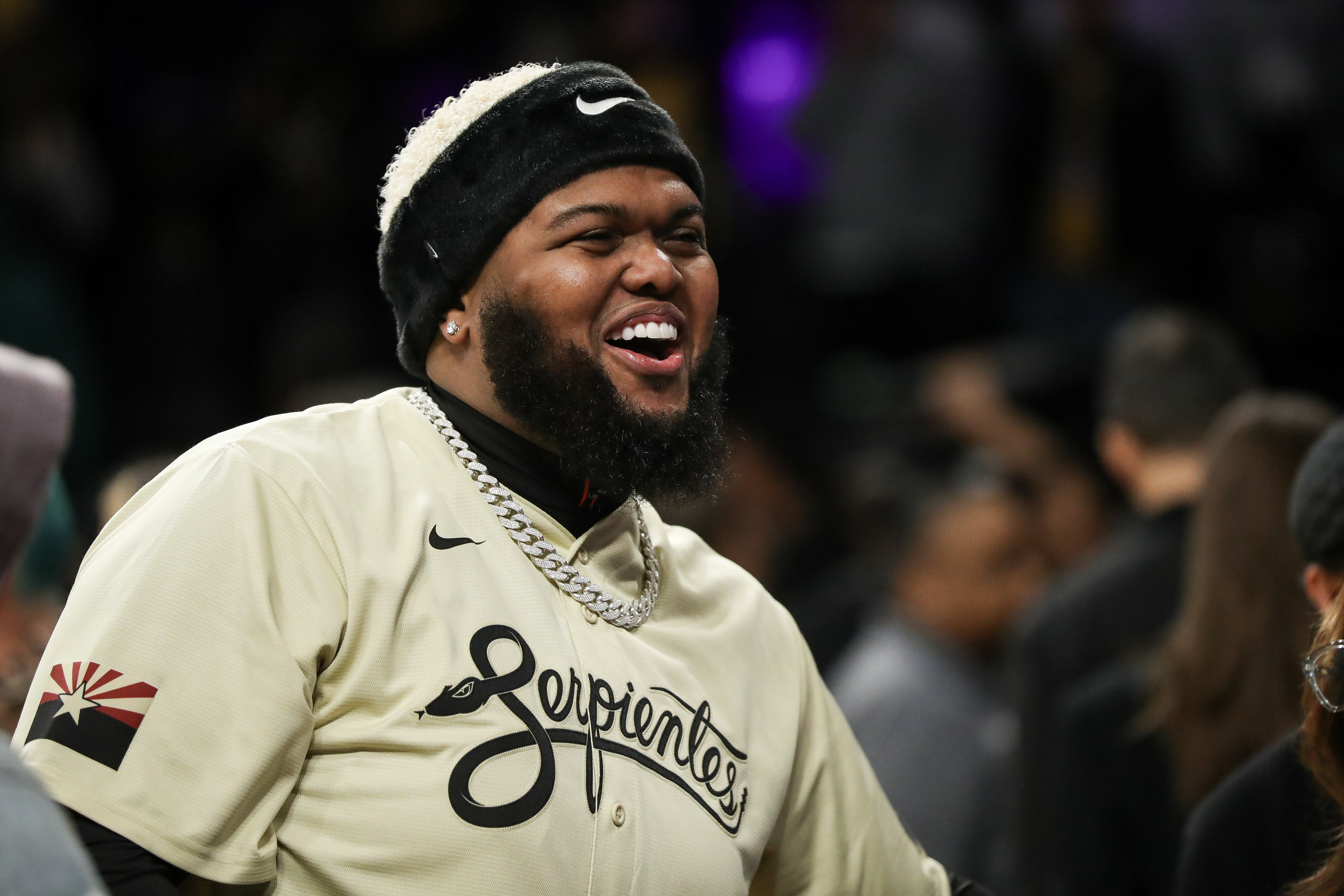 A man with a beard and black headband, wearing a beige &quot;Serpientes&quot; jersey with a silver chain necklace, is smiling widely in a crowd