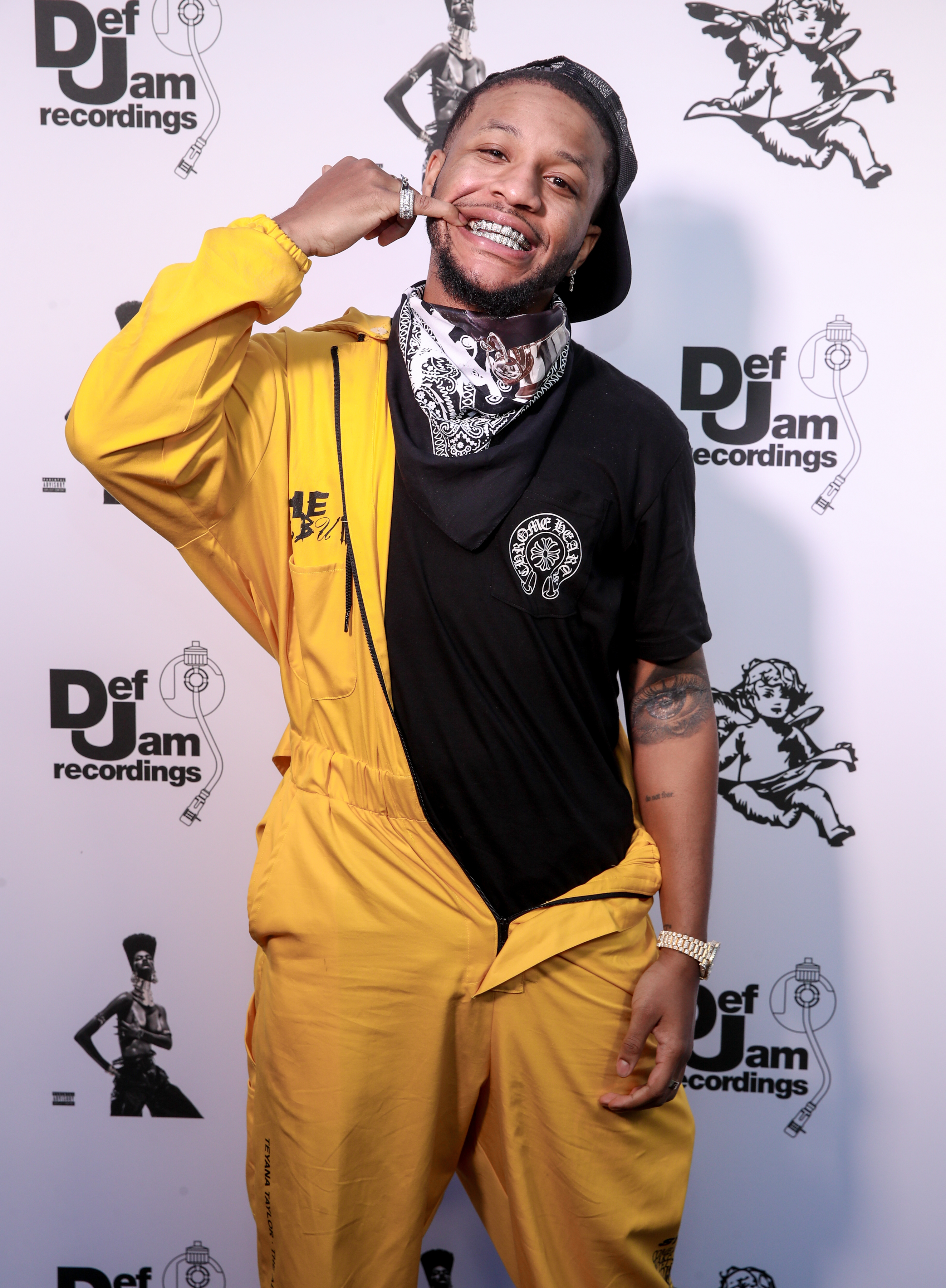 Man poses and smiles at a Def Jam Recordings event, wearing a black T-shirt, yellow jumpsuit, and a patterned bandana