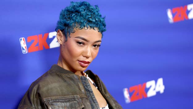 A person with short curly hair poses confidently in front of a 2K24 backdrop at a music event, wearing a casual jacket