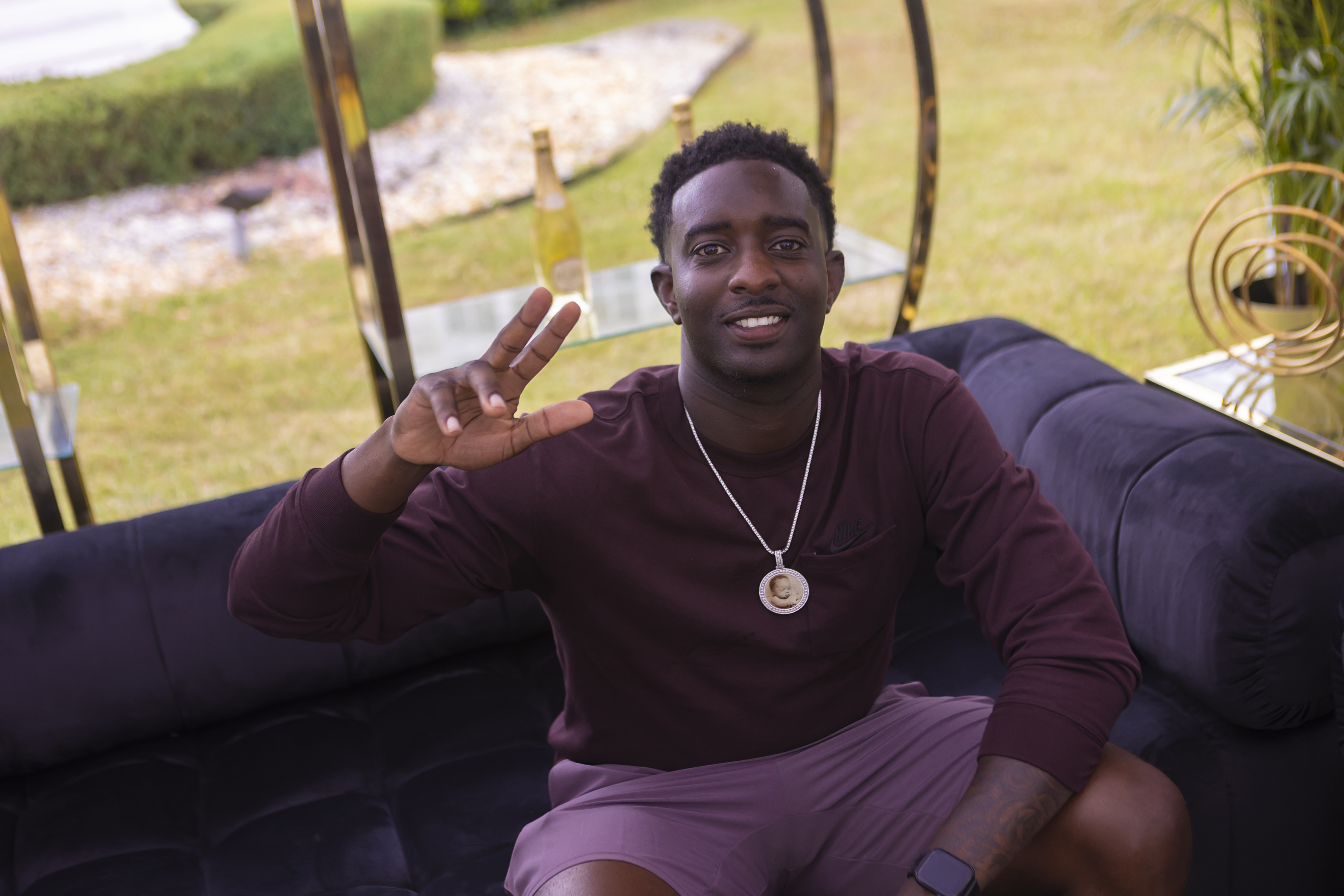 A man wearing a dark long-sleeved shirt, shorts, and a round pendant necklace is sitting on a couch outdoors, smiling and making a peace sign