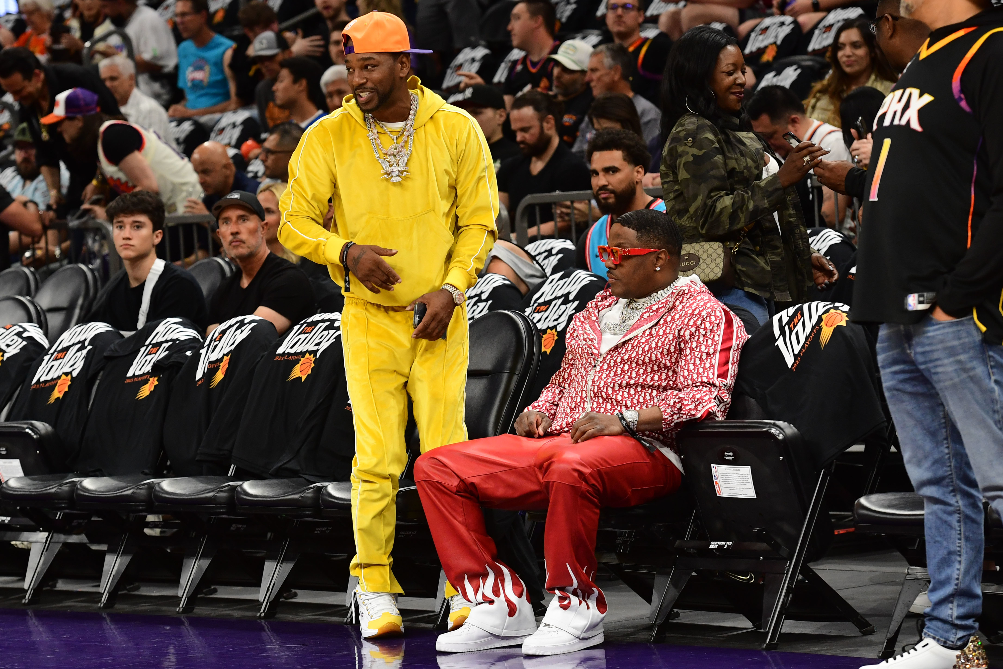 A man in a yellow tracksuit stands and talks to a seated man in a red and white ensemble with red sunglasses, courtside at a basketball game, surrounded by fans