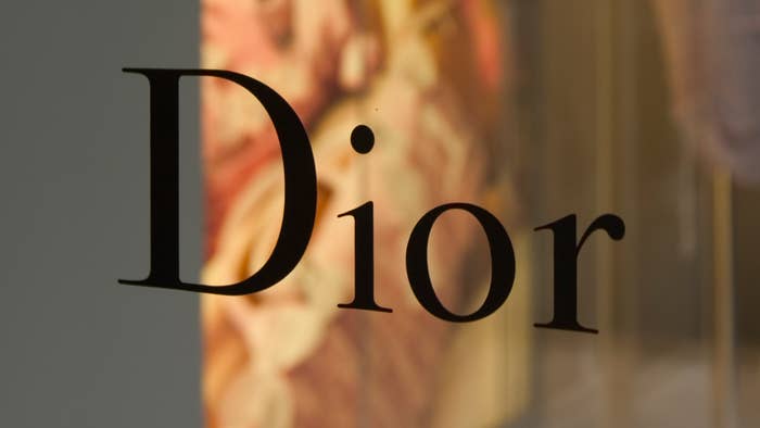 The image shows the word &quot;Dior&quot; in black letters, likely part of a store sign or advertisement. The background is blurred, featuring indistinct patterns