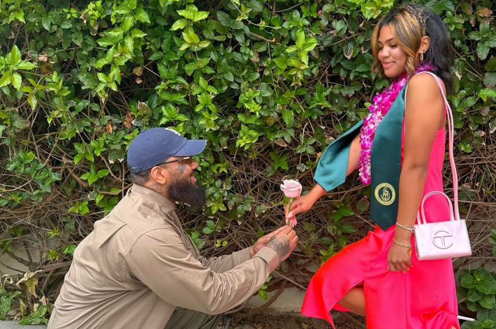 Man kneels, proposing with a flower to a woman in graduation attire with a pink purse. Woman looks at him smiling, wearing a green graduation stole