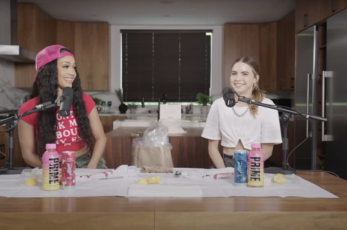 Violet Benson (in a pink hat, red top) and Elsa Majimbo (white top) are sitting at microphones in a modern kitchen, smiling and talking. Various drinks are on the table